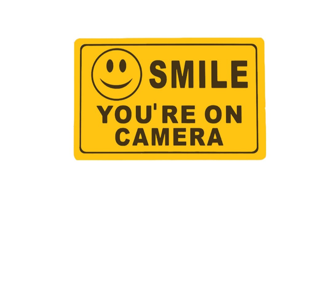 Yellow sign with smiley face and text 'SMILE YOU'RE ON CAMERA' on a white background
