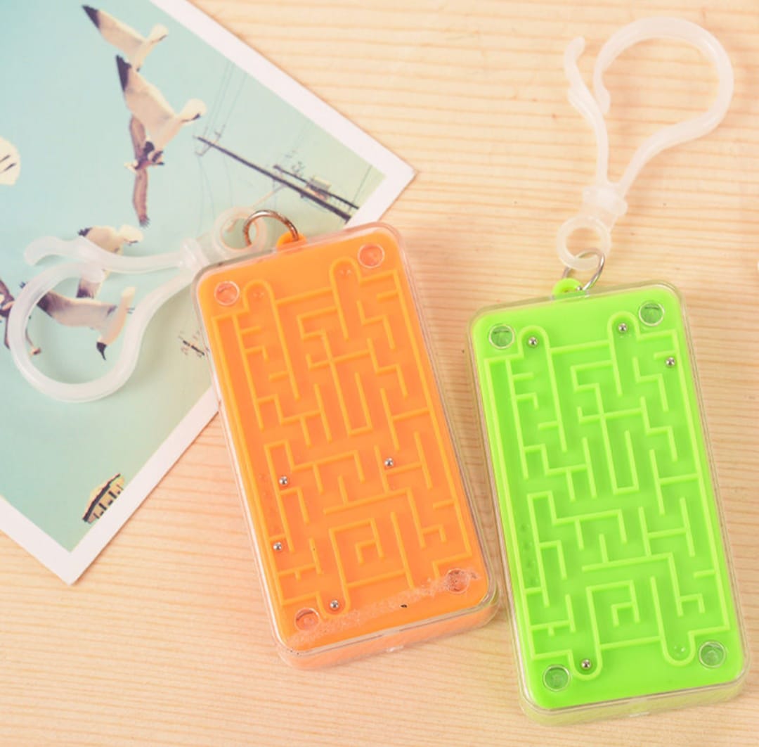 Two maze keychains, one orange and one green, on a wooden surface with a magazine in the background.